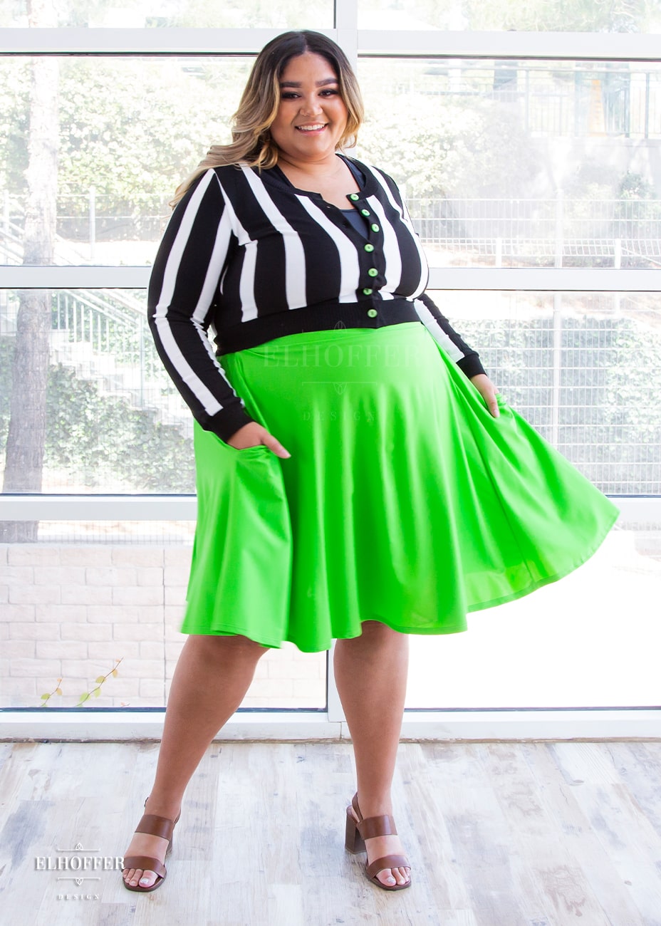 Cori, a medium skinned size 2XL model with brunette and blonde ombre hair, wears a high-waisted knee length full skirt with a fitted matching waistband encased with elastic in showtime green, a bright almost neon green. She swishes the skirt to show the movement of the fabric. She has paired it with our showtime striped cardigan.