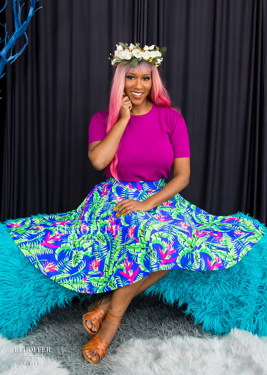 Lynsi, a dark skinned size medium model wearing a long pink ombre wig, Bernadette, a size large model with short black hair and bangs, wears a high-waisted knee length full skirt with a fitted matching waistband encased with elastic in our tropical daydream. The tropical daydream print is blue with bright green leaves and pink tropical flowers all over.