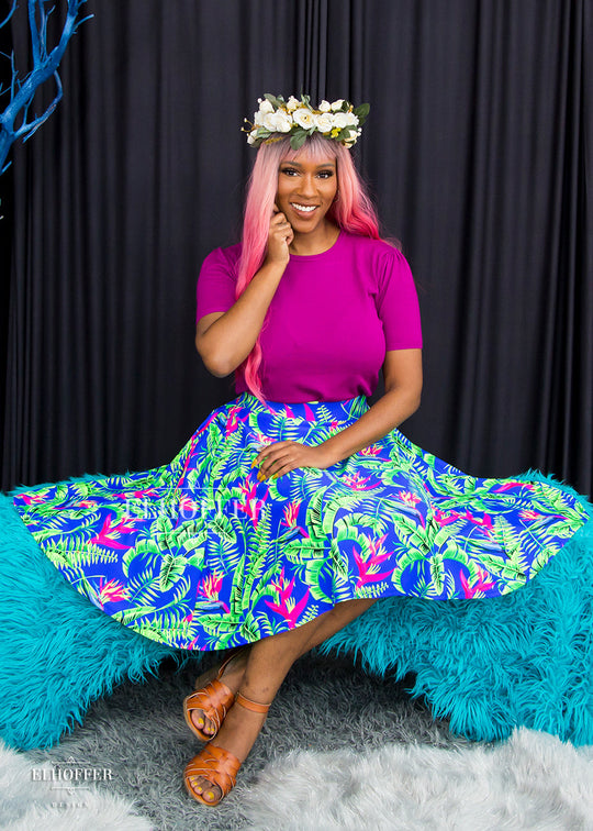 Lynsi, a dark skinned size medium model wearing a long pink ombre wig, Bernadette, a size large model with short black hair and bangs, wears a high-waisted knee length full skirt with a fitted matching waistband encased with elastic in our tropical daydream. The tropical daydream print is blue with bright green leaves and pink tropical flowers all over.