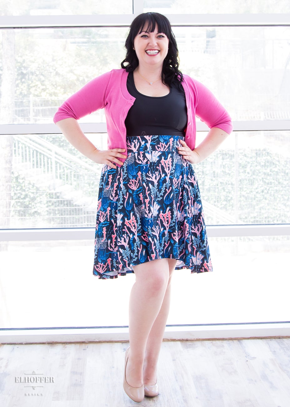 Bernadette, a fair skinned large model with short black hair and bangs, is wearing a high low skirt that hits the back of her knees with a two inch elastic covered waistband. The skirt is our barrier reef print. It is a navy based print with pink and blue coral repeated on it.