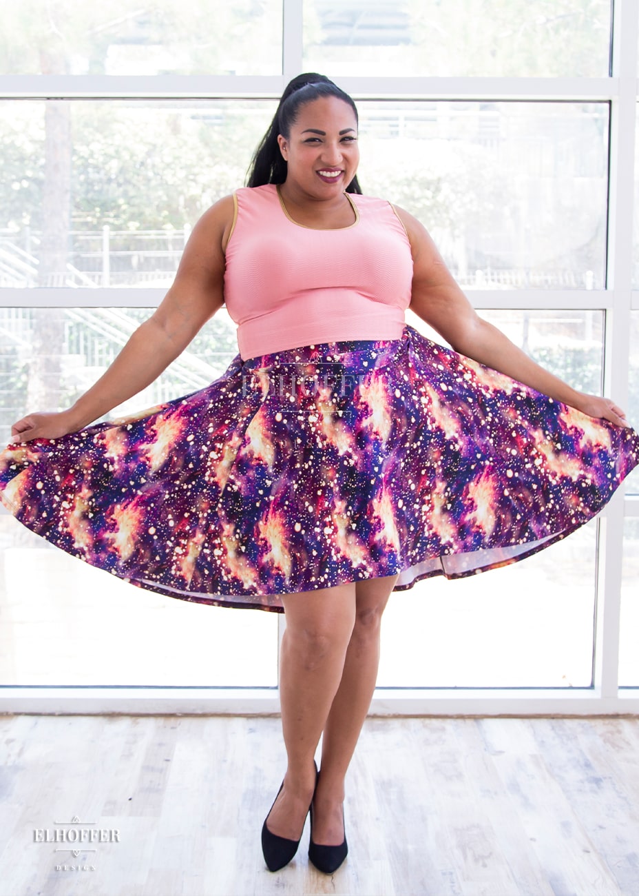 Tas, a medium dark skinned 2XL model with long dark hair, is wearing a high low skirt that hits the back of her knees with a two inch elastic covered waistband. The skirt is our bleach galaxy print. The Bleach Galaxy print is a red, purple, and pink printed galaxy pattern with light yellow splotches that appear like the fabric has been bleached all over.