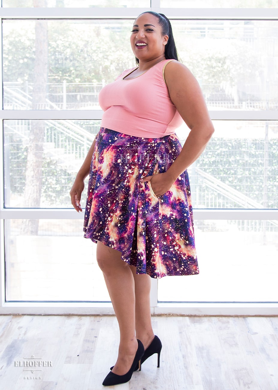 Tas, a medium dark skinned 2XL model with long dark hair, is wearing a high low skirt that hits the back of her knees with a two inch elastic covered waistband. The skirt is our bleach galaxy print. The Bleach Galaxy print is a red, purple, and pink printed galaxy pattern with light yellow splotches that appear like the fabric has been bleached all over.