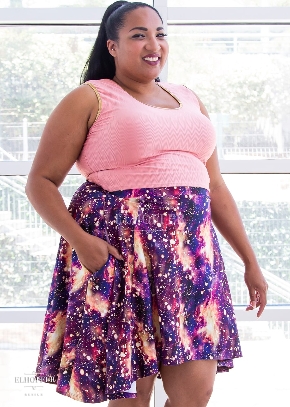 Tas, a medium dark skinned 2XL model with long dark hair, is wearing a high low skirt that hits the back of her knees with a two inch elastic covered waistband. The skirt is our bleach galaxy print. The Bleach Galaxy print is a red, purple, and pink printed galaxy pattern with light yellow splotches that appear like the fabric has been bleached all over.