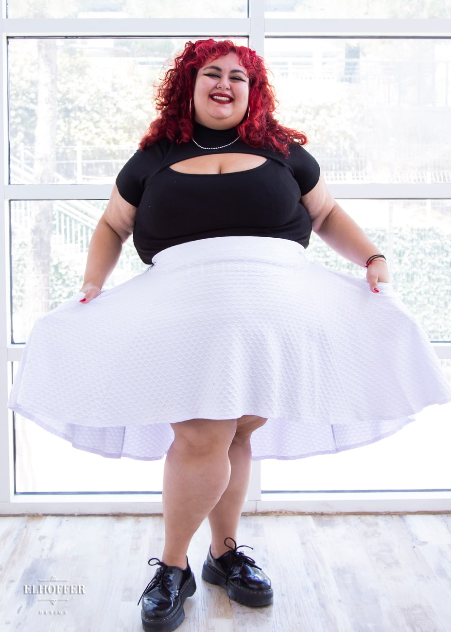 Victoria, an olive skinned size 4XL model with bright red curly hair,  is wearing a high low skirt that hits the back of her knees with a two inch elastic covered waistband. The skirt is white with a raised scale texture.