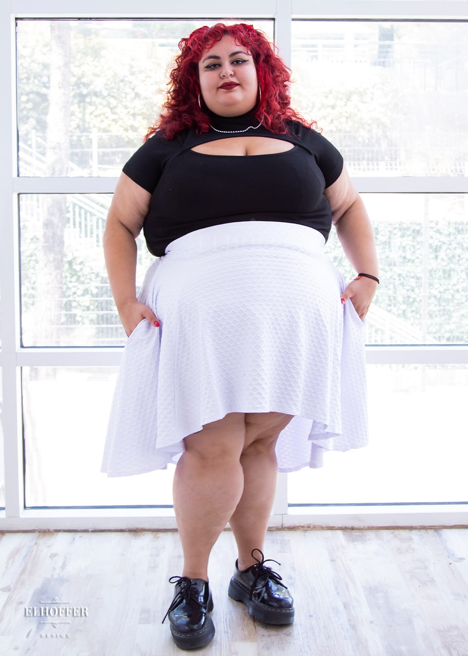 Victoria, an olive skinned size 4XL model with bright red curly hair, is wearing a high low skirt that hits the back of her knees with a two inch elastic covered waistband. The skirt is white with a raised scale texture.