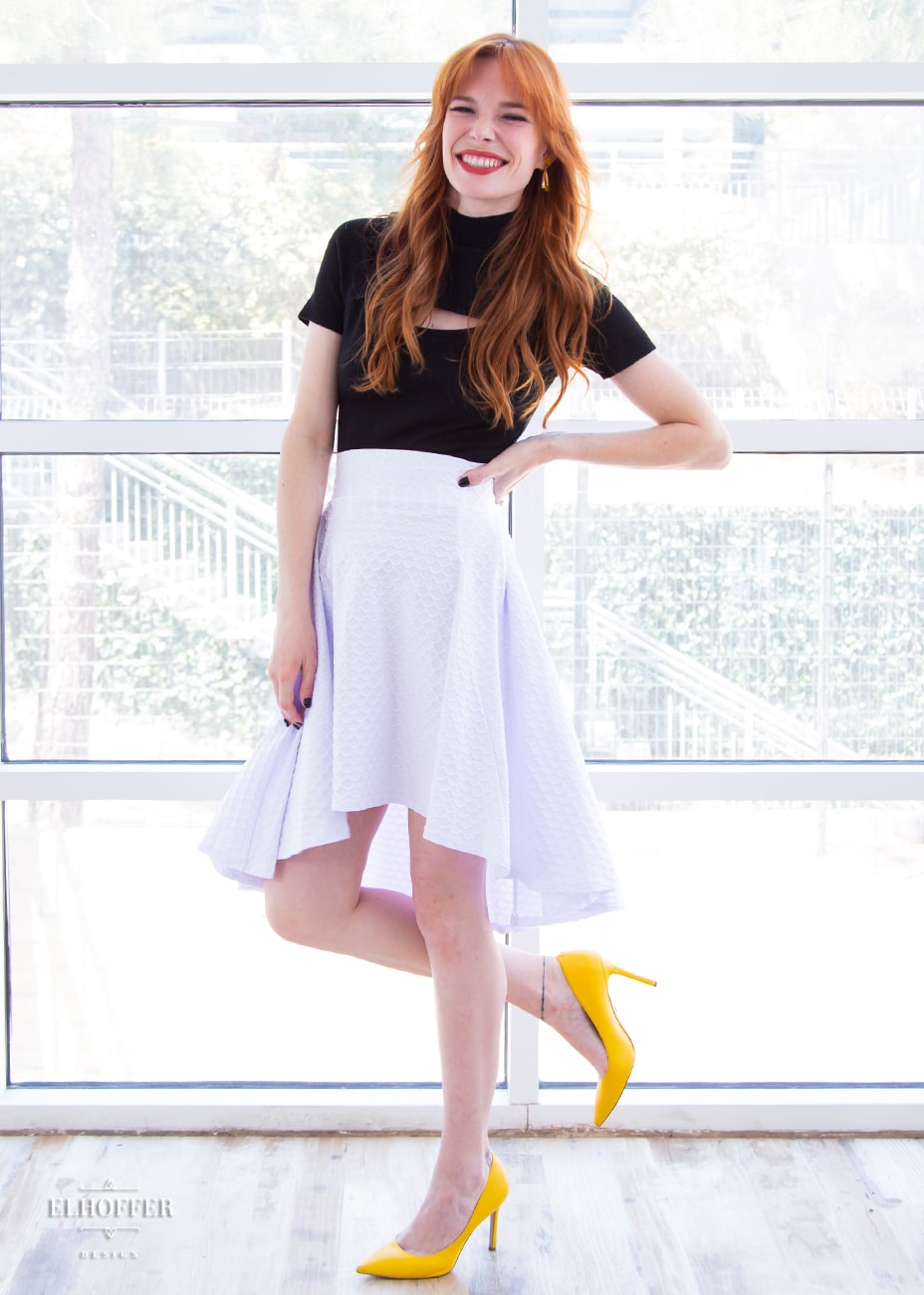 Chloe, a fair skinned XS model with red hair and bangs, is wearing a high low skirt that hits the back of her knees with a two inch elastic covered waistband. The skirt is white with a raised scale texture.