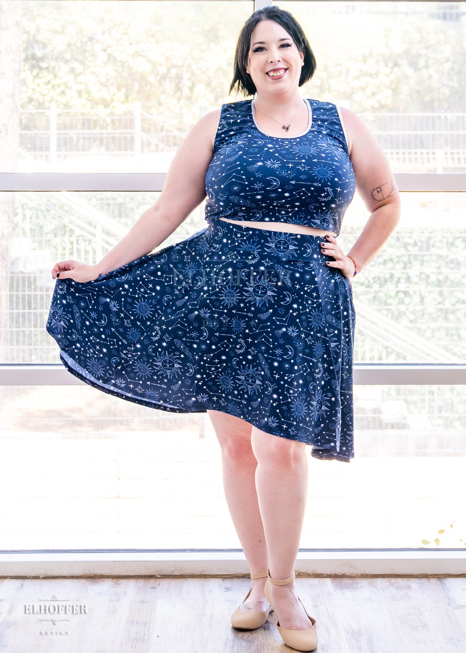 Katie Lynn is a size 2XL model with fair skin and short dark hair. She is wearing a high low skirt that hits the back of her knees with pockets and a two inch elastic covered waistband. The Star-Crossed Lovers print is a celestial inspired pattern featuring a navy base and white stars, constellations, sunds, moons, and other small details scattered across the repeated art.