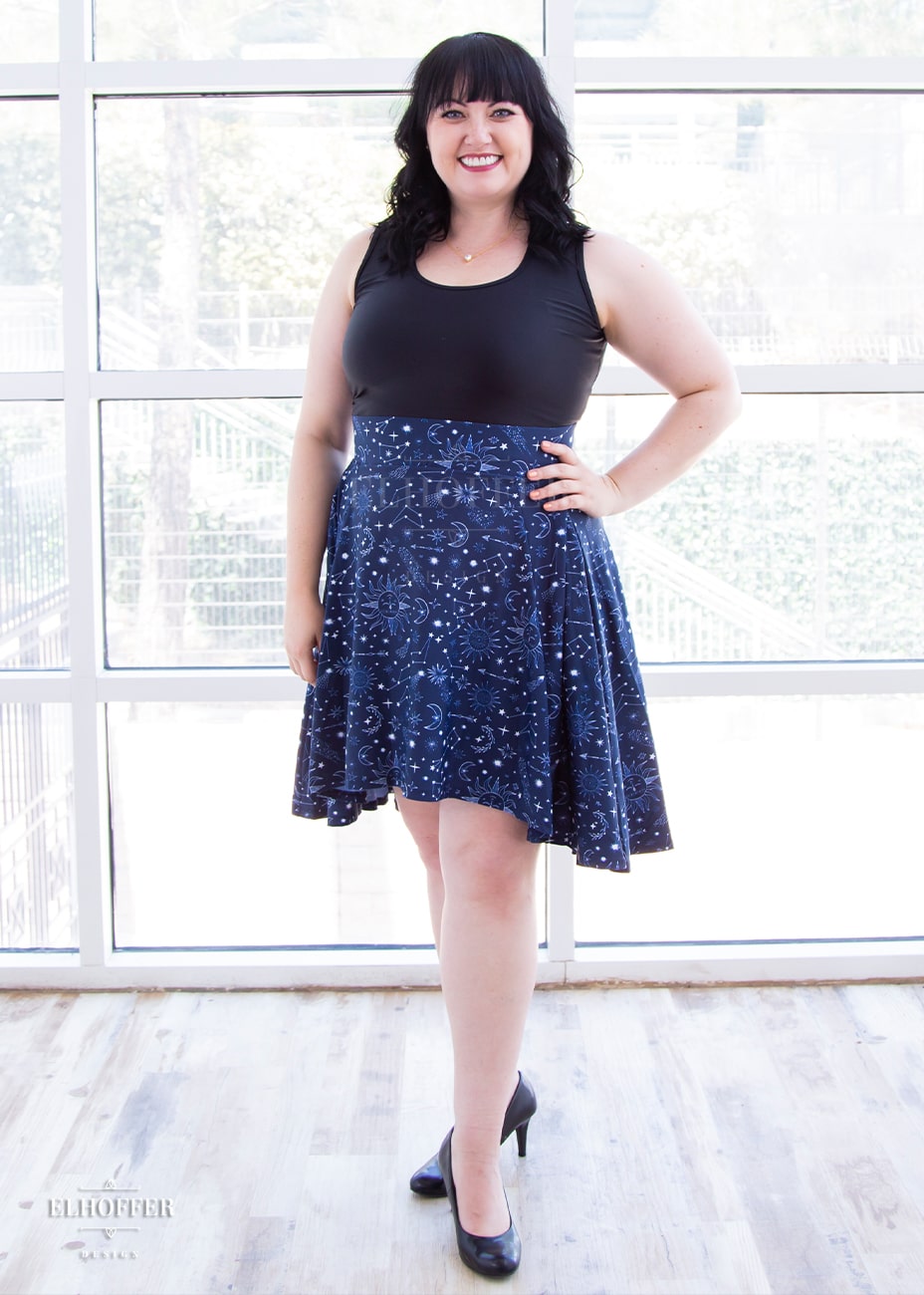 Bernadette, a fair skinned size large model with short black hair and bangs,  is wearing a high low skirt that hits the back of her knees with a two inch elastic covered waistband. The skirt is our star crossed lovers print. The Star-Crossed Lovers print is a celestial inspired pattern featuring a navy base and white stars, constellations, sunds, moons, and other small details scattered across the repeated art.