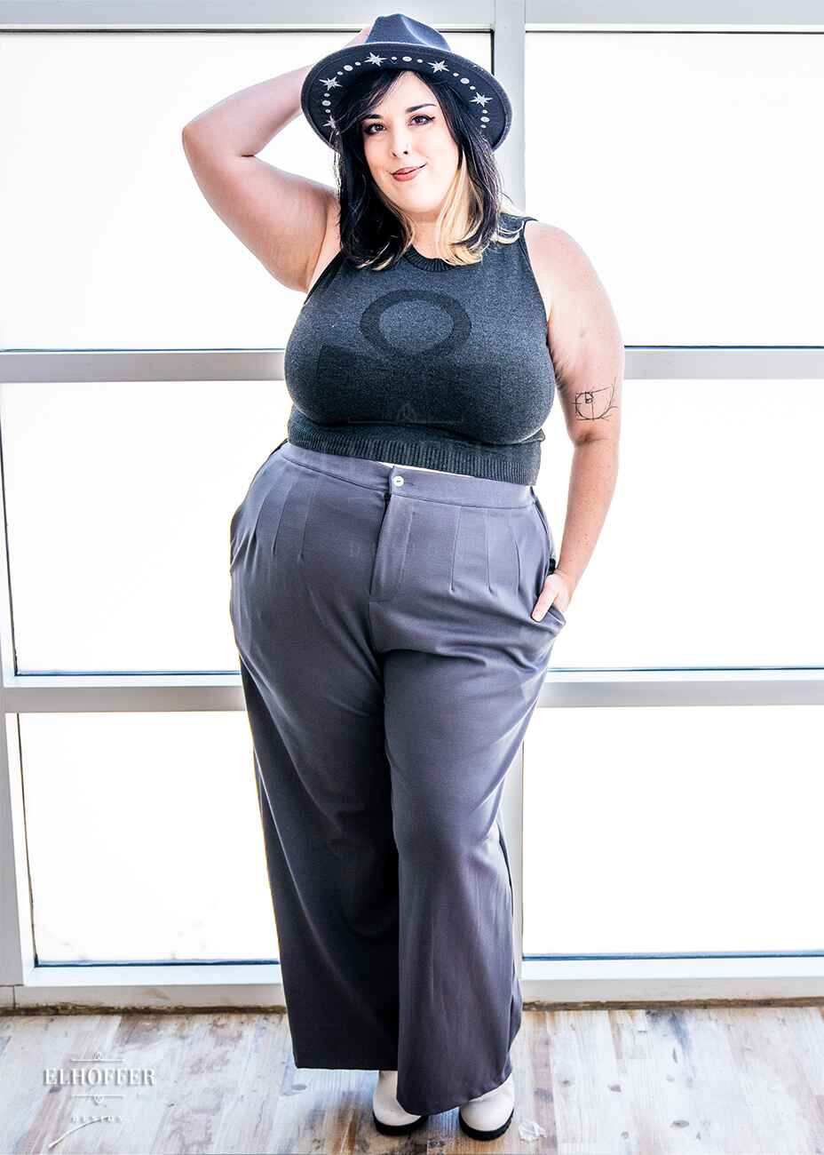 Katie Lynn, a fair skinned 2xl model with shoulder length black and blonde hair is wearing a dark grey sleeveless knit crop top with a large subtle ankh design on the front.  She paired the crop top with grey pleated trousers.