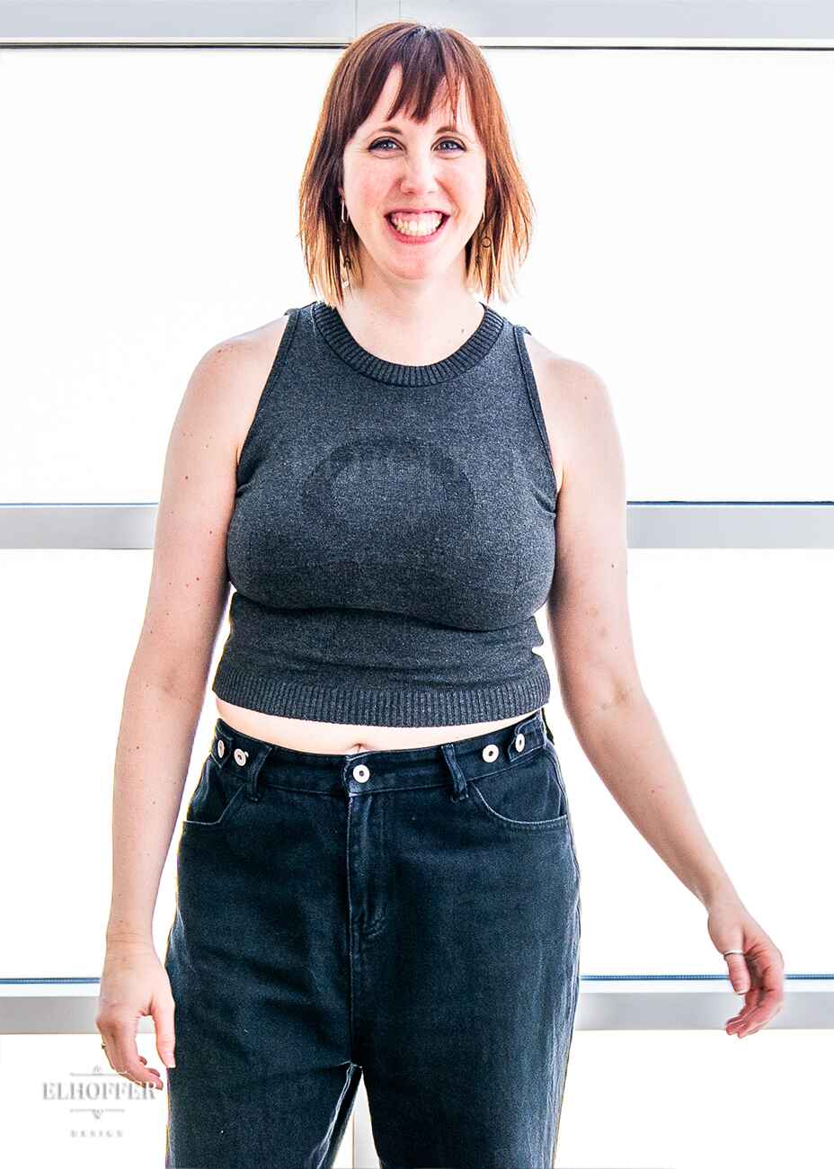 Jes, a fair skinned S model with short red hair with bangs, is caught mid laugh while wearing a dark grey sleeveless knit crop top with a large subtle ankh design on the front.