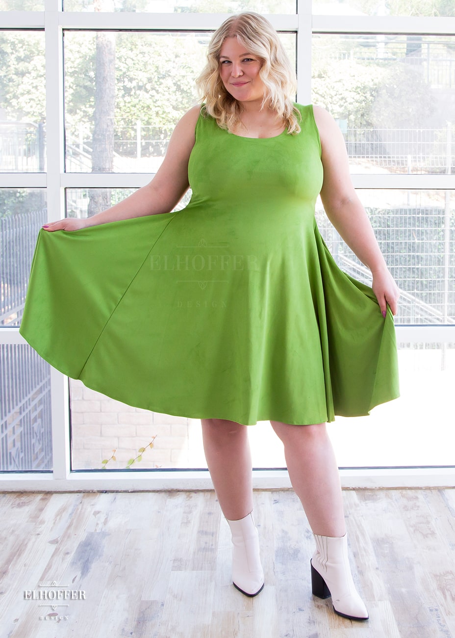 Sarah, a fair skinned size XL model with long blonde hair, wears a light green suede knee length dress with fitted torso and full skirt. She holds out the side of the dress to show off the generous amount of fabric.