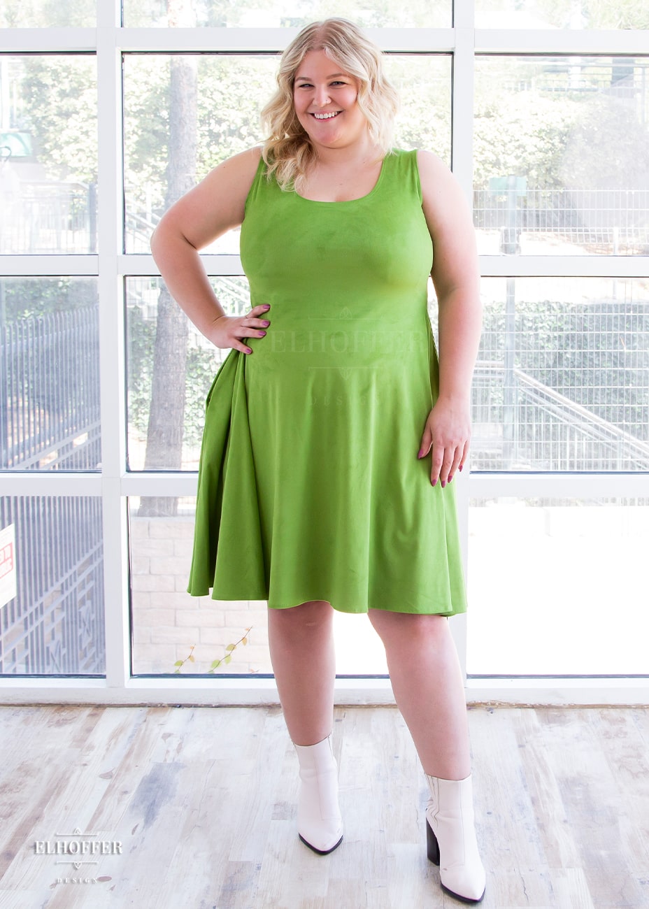 Sarah, a fair skinned size XL model with long blonde hair, wears  a light green suede knee length dress with fitted torso and full skirt. Her hand is on her waist to show off the fitted torso.