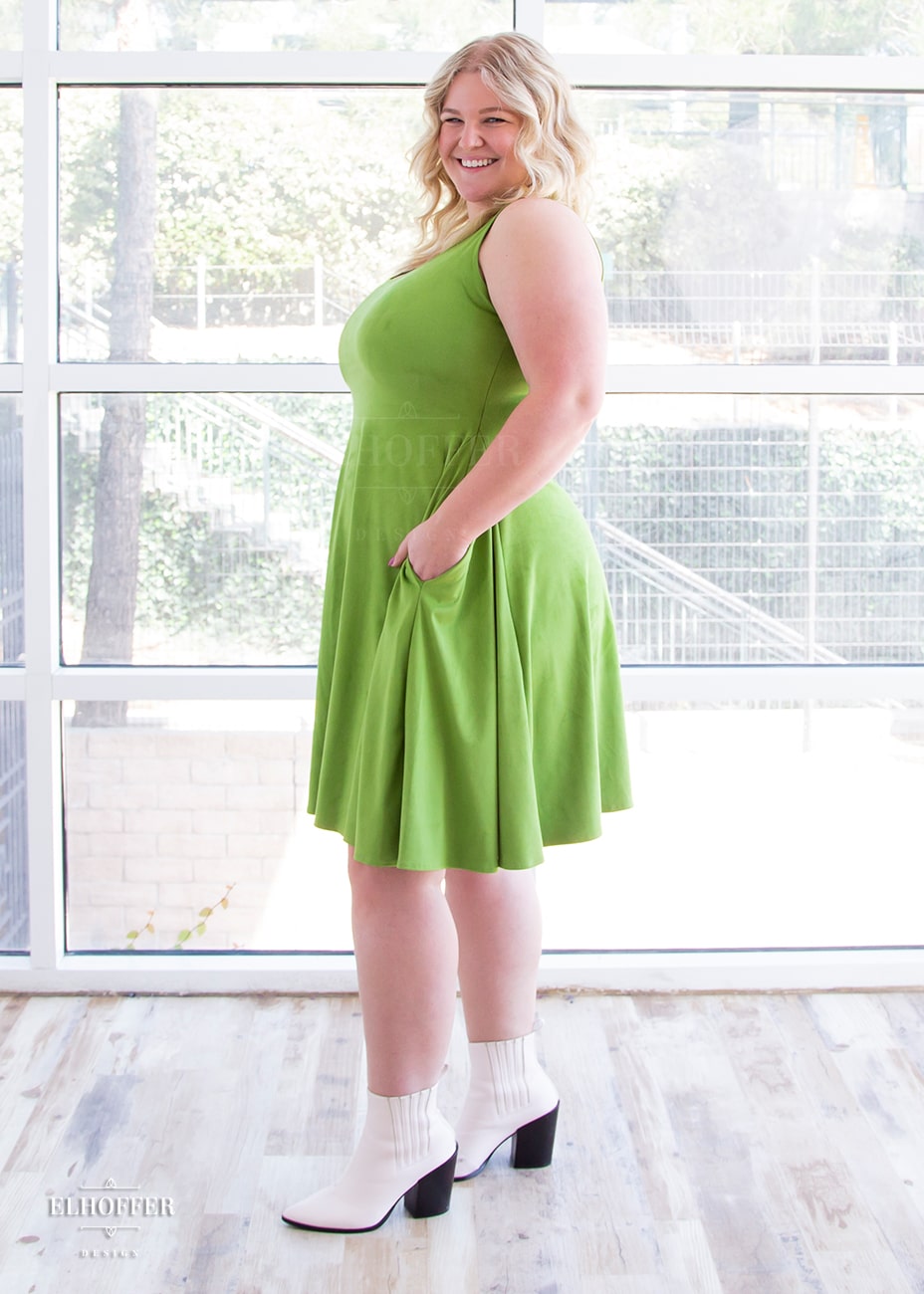 Sarah, a fair skinned size XL model with long blonde hair, wears a light green suede knee length dress with fitted torso and full skirt. She stands to the side and has her hand in the generous pocket.