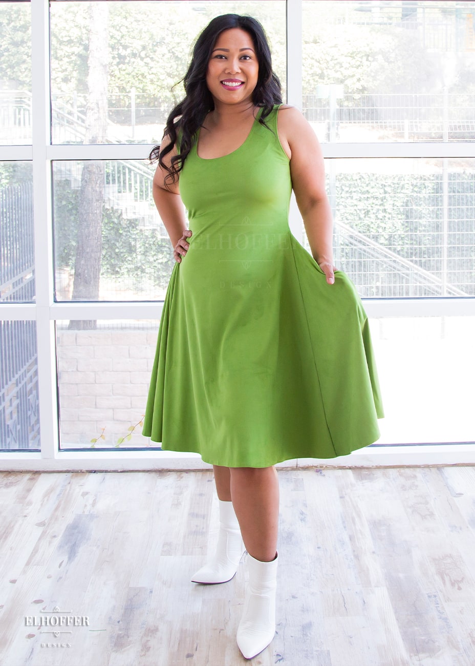 Alyssa, a medium skinned size medium model with long dark hair, wears a light green suede knee length dress with fitted torso and full skirt. She holds out one side to show off the pockets.