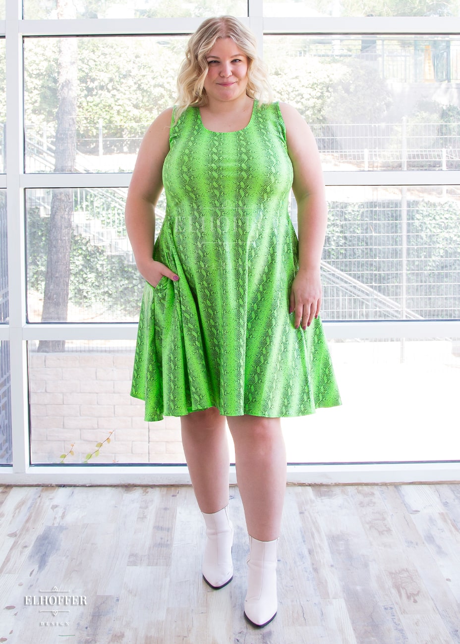 Sarah, a fair skinned size XL model with long blonde hair, wears a sleeveless knee length dress that is fitted in the torso and full in the skirt. The print is croki green, a lime green snake print. She has her hand in a generous pocket.