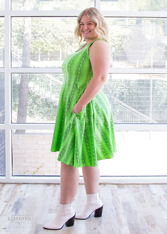 Sarah, a fair skinned size XL model with long blonde hair, wears a sleeveless knee length dress that is fitted in the torso and full in the skirt. The print is croki green, a lime green snake print. She faces to the side and has her hand in a generous pocket.