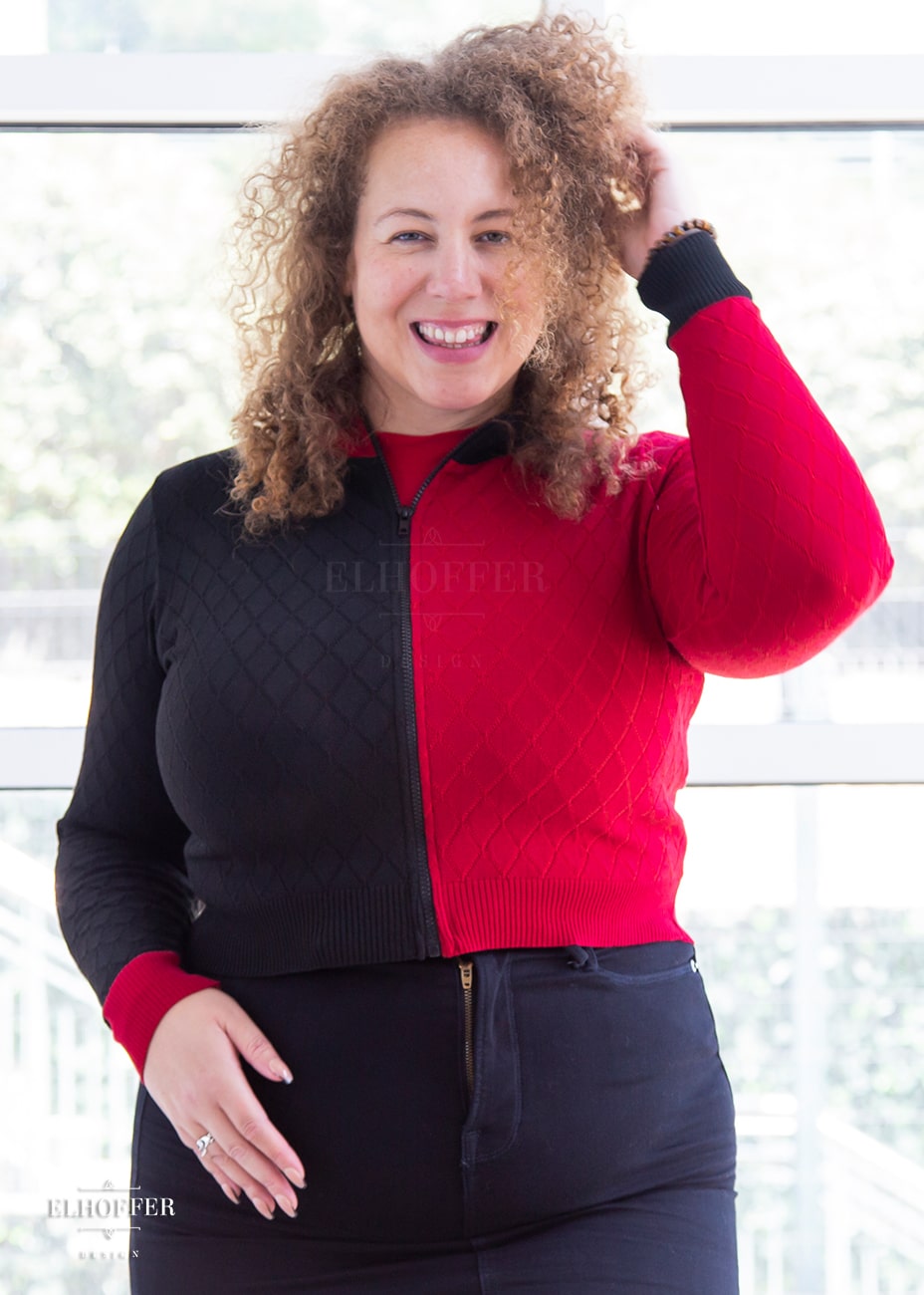 Anastasia, a curly haired, medium-skinned size XL model, is wearing our Kwinn zip up cardigan. It is a zip up cardigan with fitted long sleeves and a high neckline. The knit body has a diamond pattern, one side is black, the other red, with opposite colored cuffs.