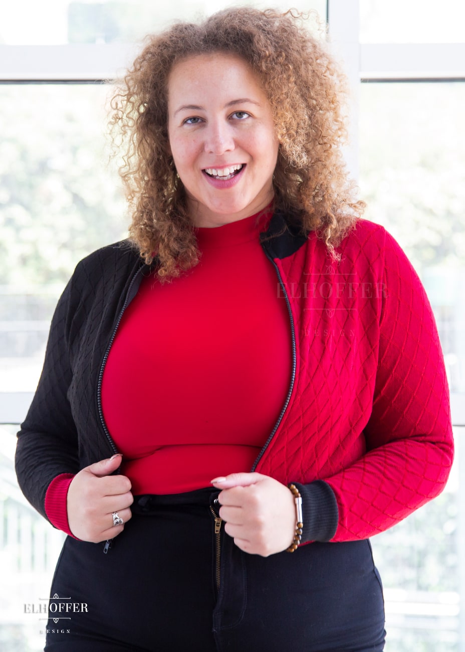 Anastasia, a curly haired, medium-skinned size XL model, is wearing our Kwinn zip up cardigan. It is a zip up cardigan with fitted long sleeves and a high neckline. The knit body has a diamond pattern, one side is black, the other red, with opposite colored cuffs.