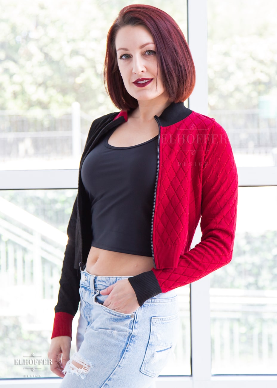 Natalie, a size small fair skinned model with short red hair, is wearing our Kwinn zip up cardigan. It is a zip up cardigan with fitted long sleeves and a high neckline. The knit body has a diamond pattern, one side is black, the other red, with opposite colored cuffs.