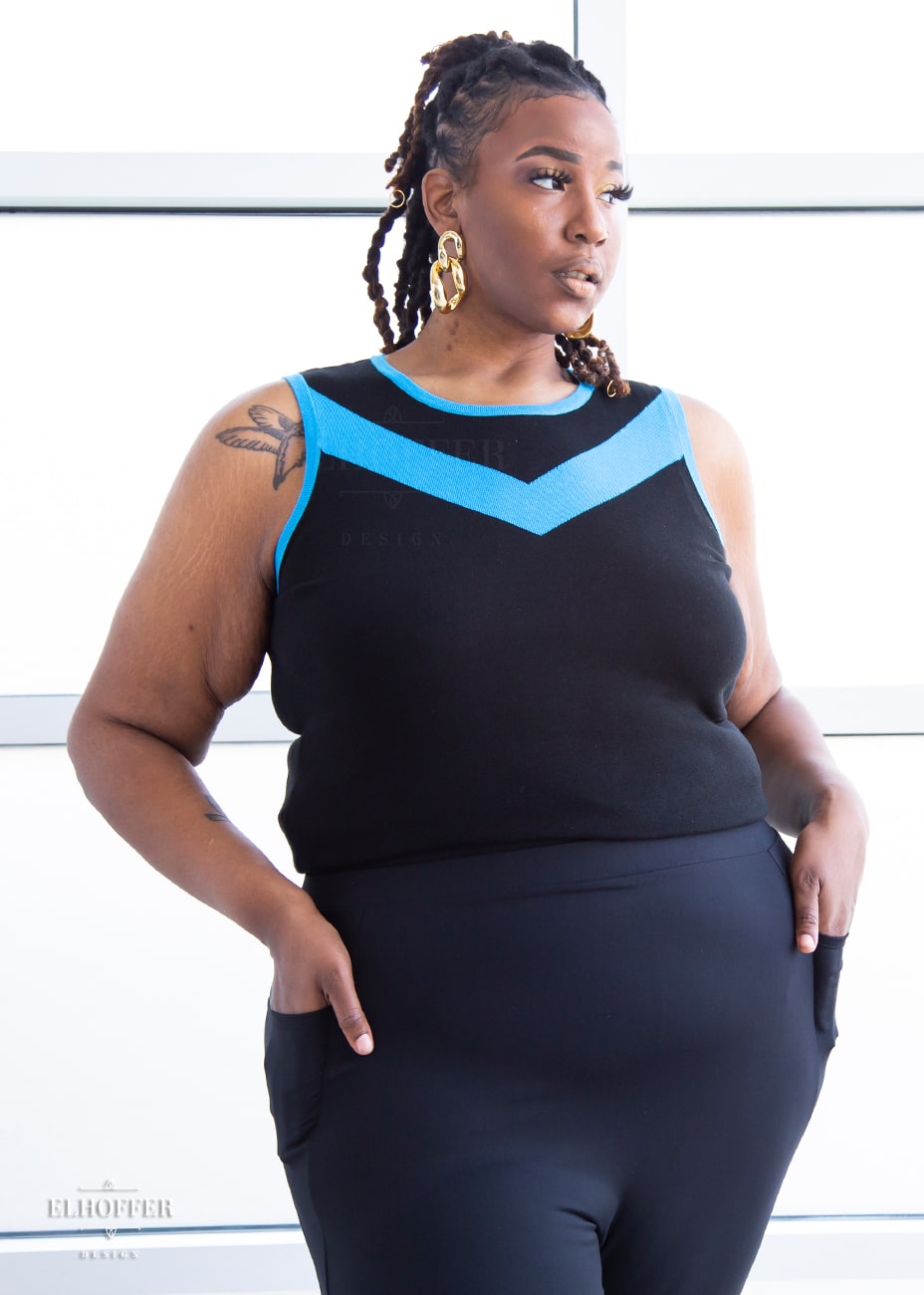 Myjah, a medium dark skinned 3xl model with shoulder length braids, is wearing a light weight sleeveless knit top.  The body of the top is mainly black with a bright teal chevron design across the chest and matching teal binding around the neckline and armholes.