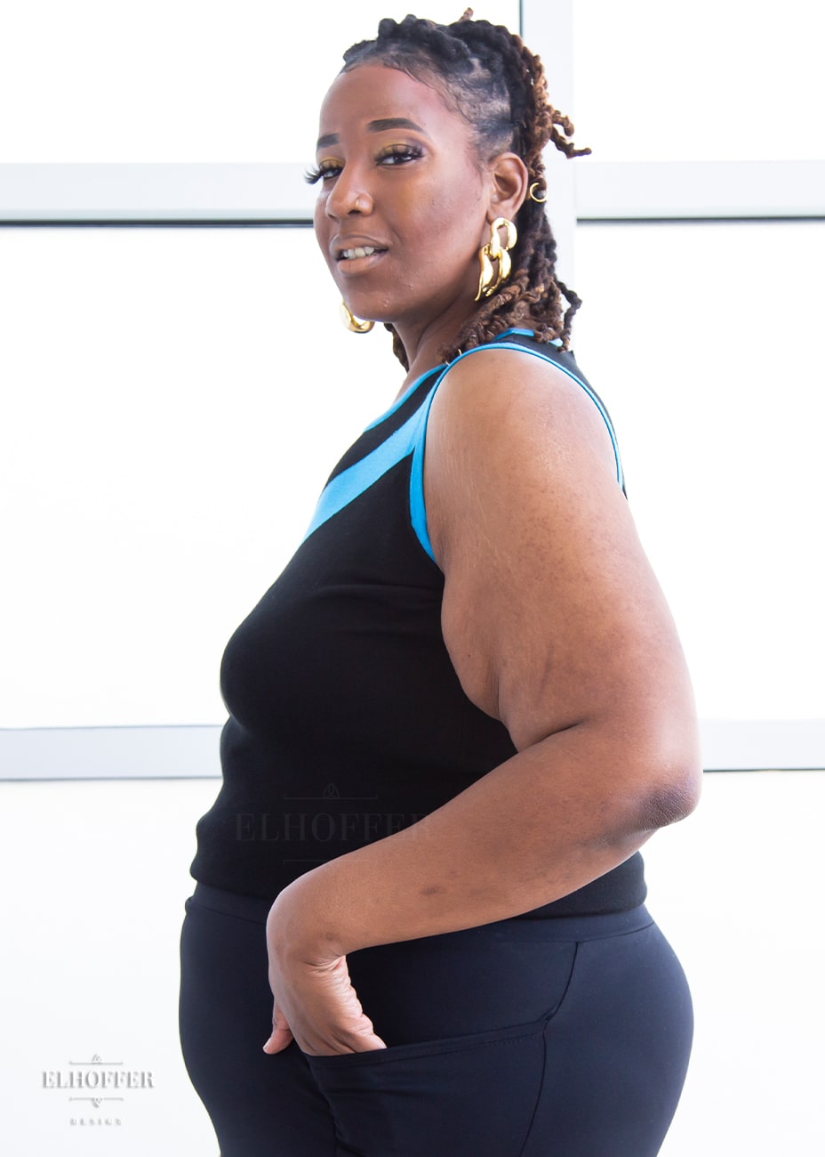 Side view of Myjah, a medium dark skinned 3xl model with shoulder length braids, wearing a light weight sleeveless knit top.  The body of the top is mainly black with a bright teal chevron design across the chest and matching teal binding around the neckline and armholes.
