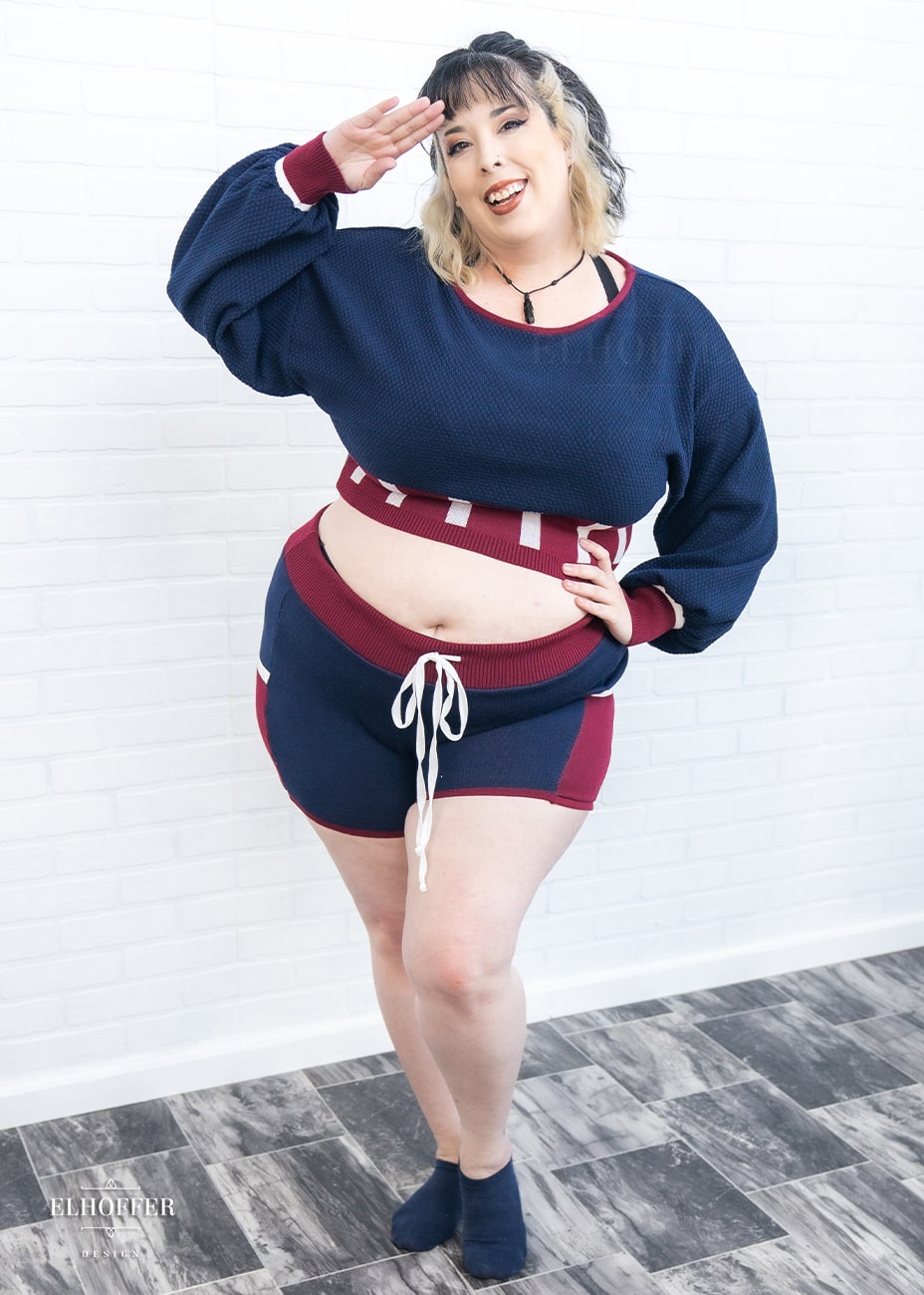 Katie Lynn, a size 2XL fair skinned model with black and blonde hair, is wearing the red, white and blue knit oversized crop top with bishop sleeves. The sleeves and top of the crop are blue with a waffle knit, the waist is vertical red and white stripes. The cuffs are mainly red with a white detail. There is also a red detail at the neckline.