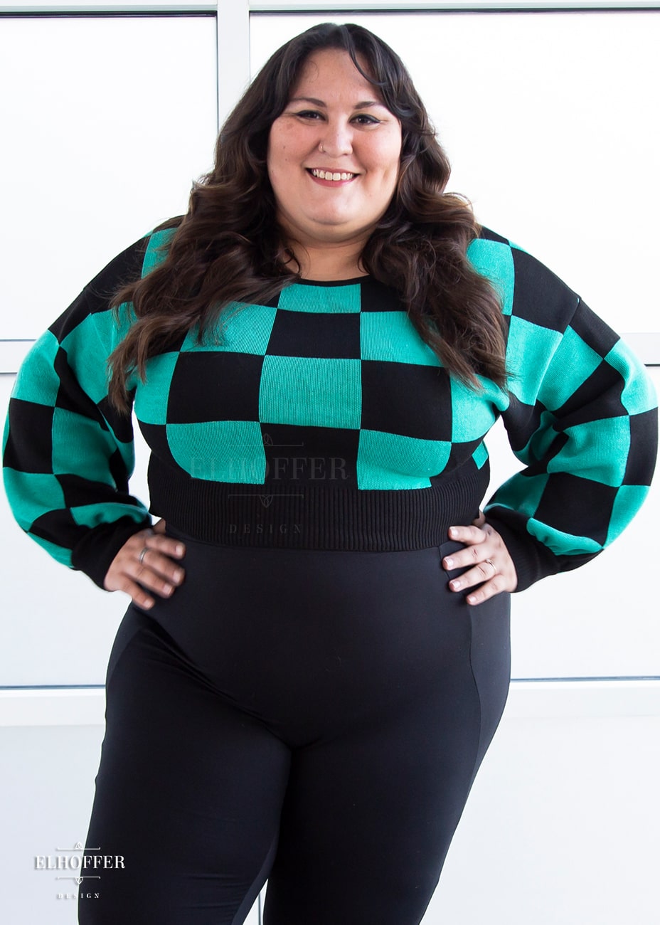 Alysia, a sun kissed skin 2xl model with long dark brown curly hair, is smiling while wearing a cropped oversize sweater with a black and green chessboard pattern and long billowing sleeves.  She paired the sweater with black leggings.
