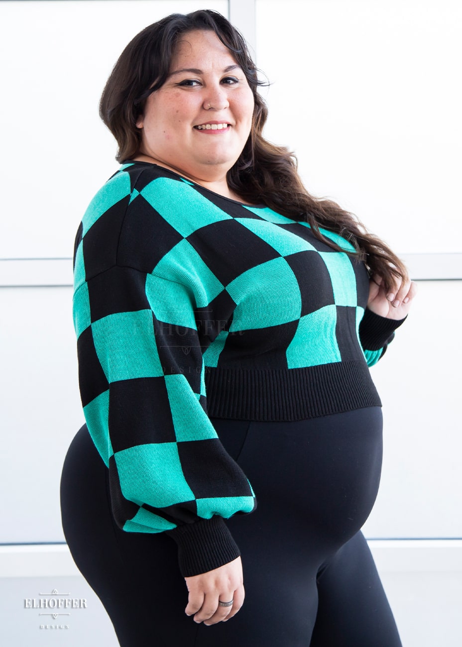 Alysia, a sun kissed skin 2xl model with long dark brown curly hair, is smiling while wearing a cropped oversize sweater with a black and green chessboard pattern and long billowing sleeves.  She paired the sweater with black leggings.