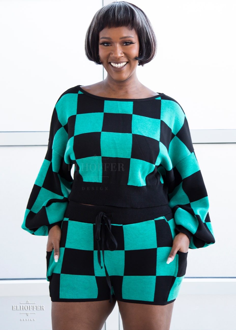 Lynsi, a medium dark skinned M model with short black and white hair, is smiling while wearing the XL/2XL sample of a cropped oversize sweater with black and green chessboard pattern and long billowing sleeves. She would normally wear the M/L. She paired them with matching shorts. Note: the matching shorts are a sample and not included nor available at this time.