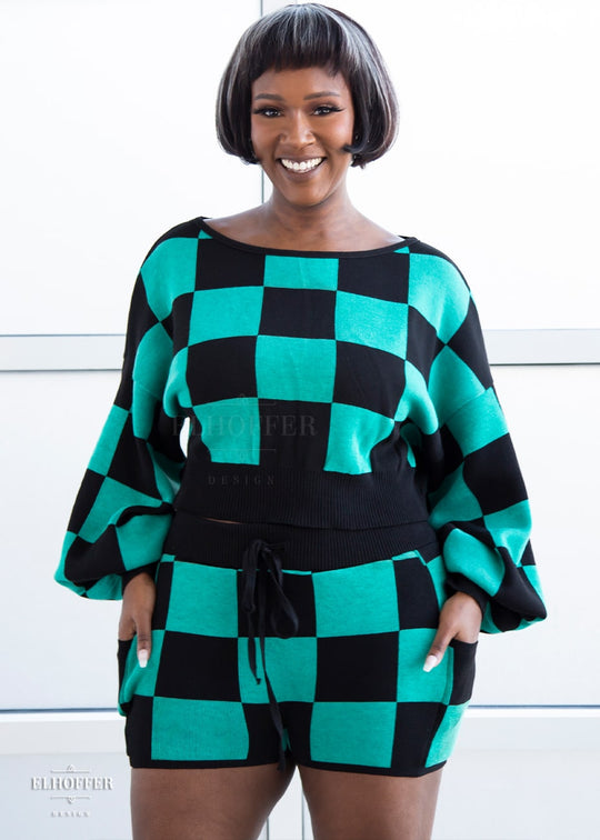 Lynsi, a medium dark skinned M model with short black and white hair, is smiling while wearing the XL/2XL sample of a cropped oversize sweater with black and green chessboard pattern and long billowing sleeves. She would normally wear the M/L. She paired them with matching shorts. Note: the matching shorts are a sample and not included nor available at this time.