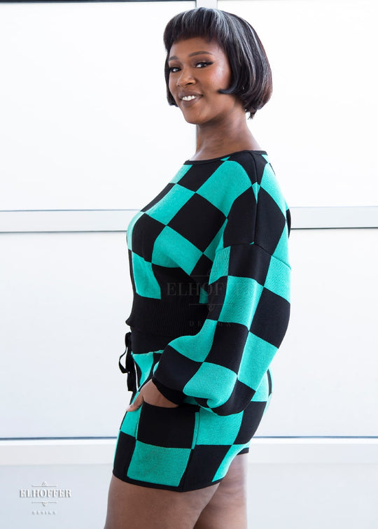 Lynsi, a medium dark skinned M model with short black and white hair, is smiling while wearing the XL/2XL sample of a cropped oversize sweater with black and green chessboard pattern and long billowing sleeves. She would normally wear the M/L. She paired them with matching shorts. Note: the matching shorts are a sample and not included nor available at this time.