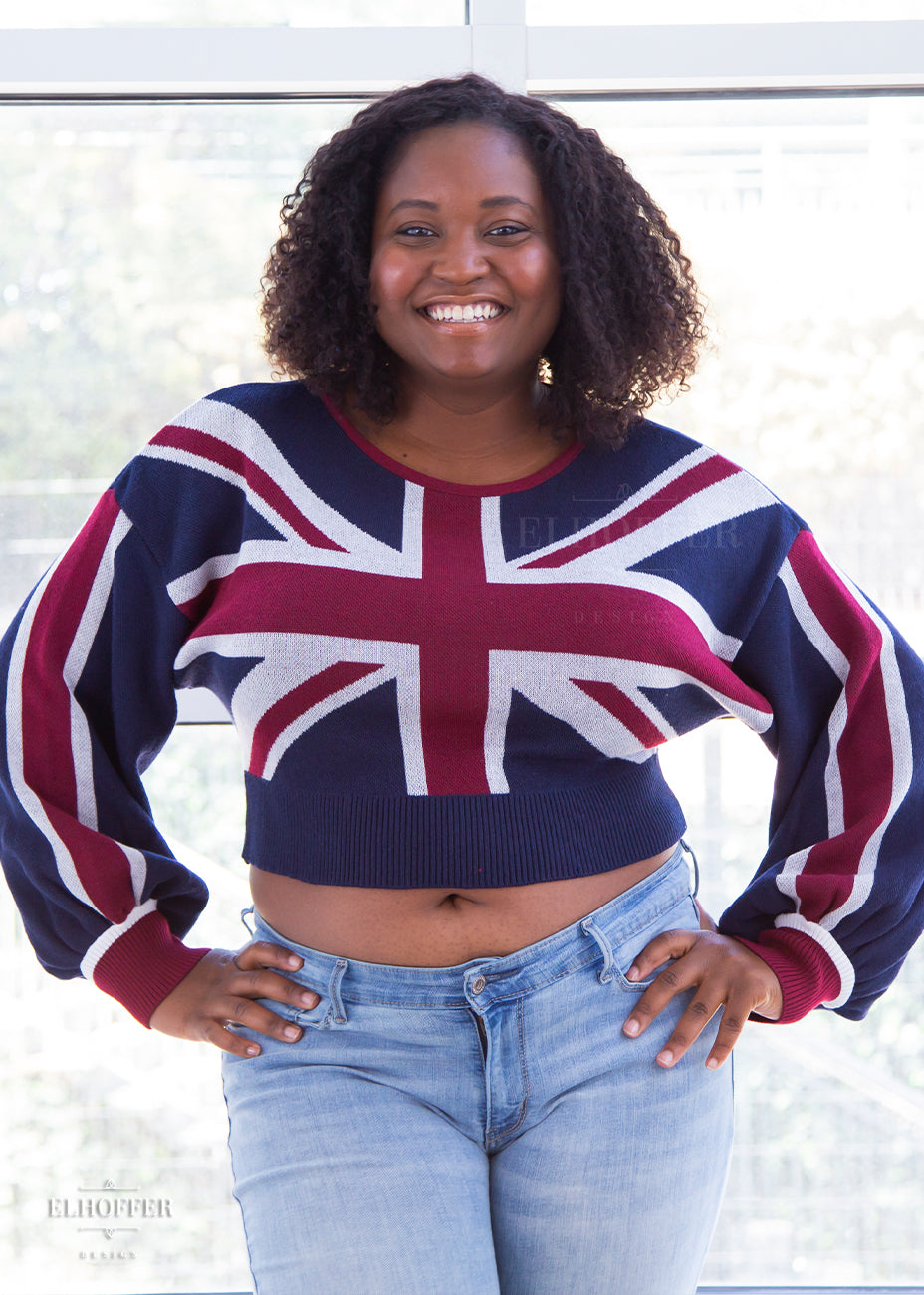 Maydelle, a medium dark skinned size XL model with shoulder length curly brown hair, is wearing an oversized drop shoulder bishop sleeve crop top emblazoned with the union jack.