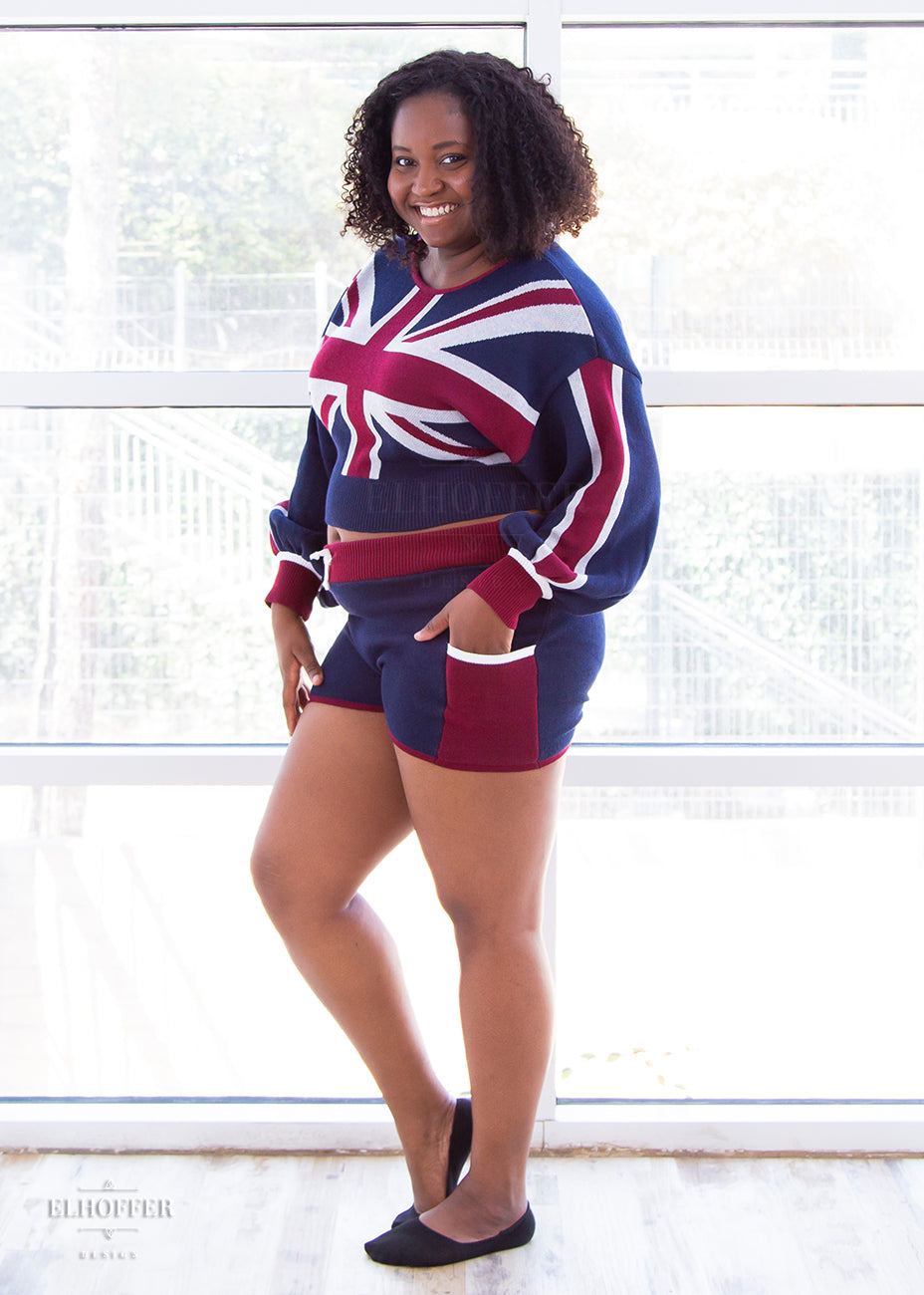 Maydelle, a medium dark skinned XL model with short curly brown hair, is wearing the red, white and blue knit oversized crop top with bishop sleeves. The top has a union jack across the chest and continuing into the sleeves. The cuffs are red with a white detail. There is also a red detail at the neckline.