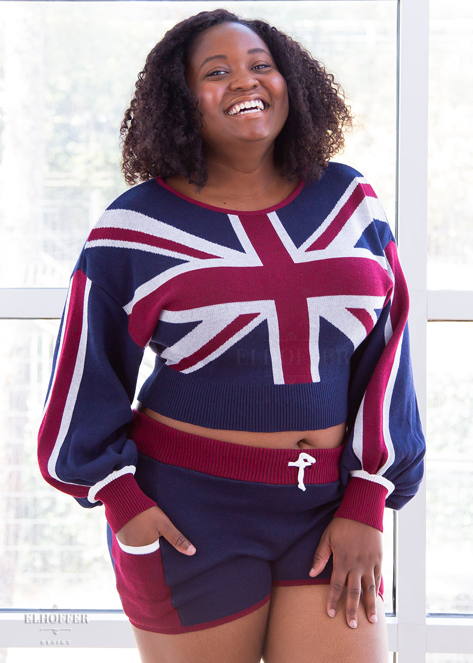 Maydelle, a medium dark skinned XL model with short curly brown hair, is wearing the red, white and blue knit oversized crop top with bishop sleeves. The top has a union jack across the chest and continuing into the sleeves. The cuffs are red with a white detail. There is also a red detail at the neckline.