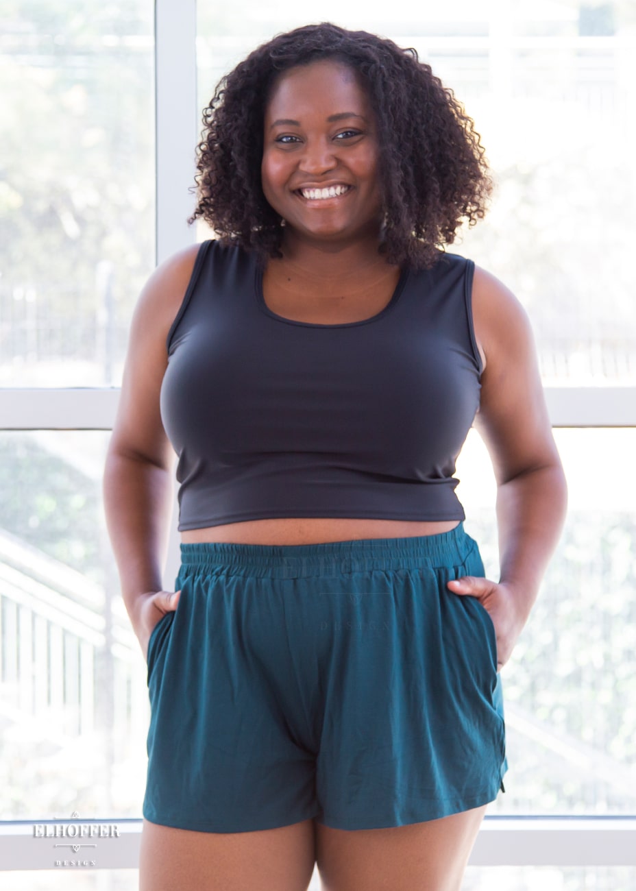 Maydelle, a medium dark skinned size XL model with shoulder length curly brown hair is wearing a pair of seamless front, medium rise, loose fit hunter green shorts with side pockets.
