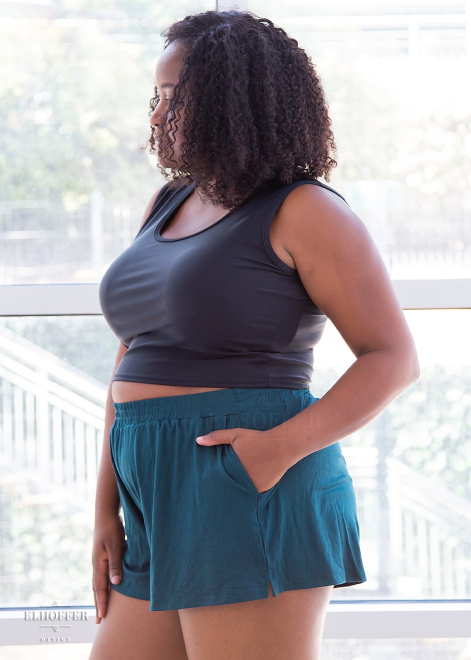 Maydelle, a medium dark skinned size XL model with shoulder length curly brown hair is wearing a pair of seamless front, medium rise, loose fit hunter green shorts with side pockets.