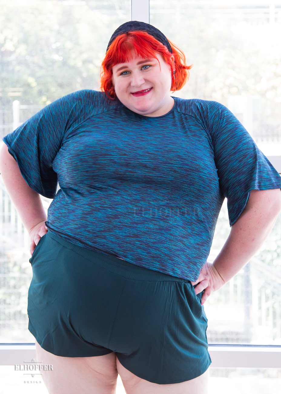 Logan, a fair skinned size 3XL model with short bright orange hair and bangs, is wearing a pullover gathered raglan sleeve top with short bell sleeves, in a heathered blue.