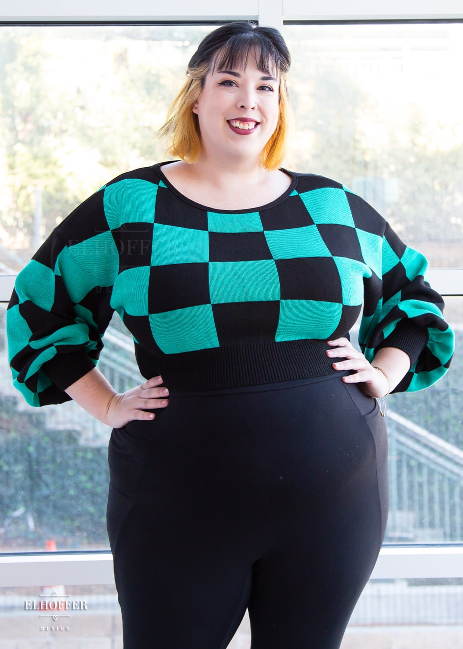 Katie Lynn, a fair skinned 2xl model with short black and blonde hair, is smiling while wearing a cropped oversize sweater with a black and green chessboard pattern and long billowing sleeves. She paired the sweater with black leggings.