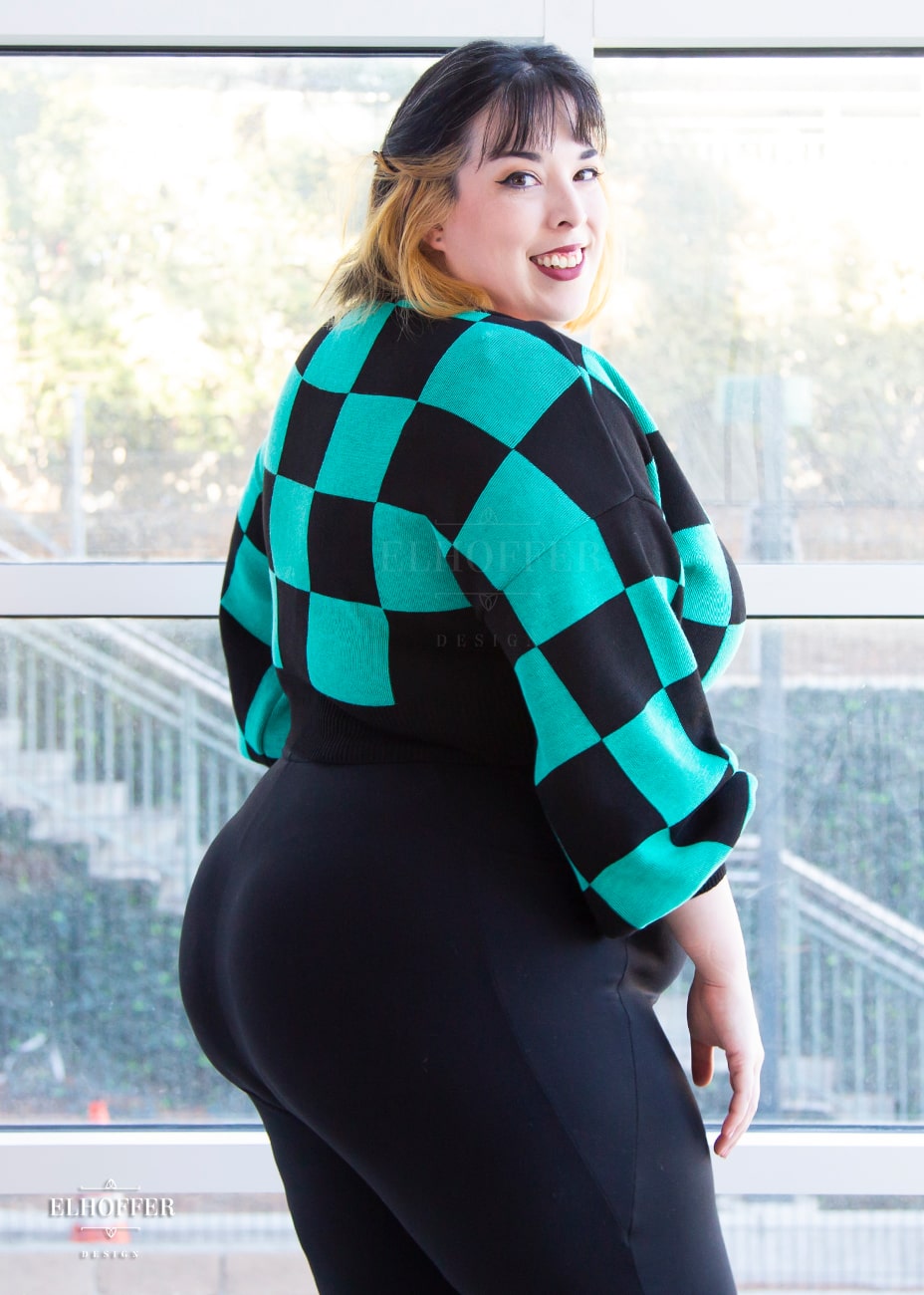 Katie Lynn, a fair skinned 2xl model with short black and blonde hair, is smiling while wearing a cropped oversize sweater with a black and green chessboard pattern and long billowing sleeves. She paired the sweater with black leggings.