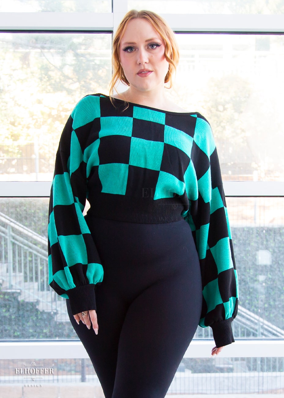 Kelsey, a fair skinned S model with strawberry blonde hair, is smiling while wearing the XL/2XL sample of a cropped oversized sweater with a black and green chessboard pattern and long billowing sleeves.  She would normally wear the XS/S.  She paired the sweater with black leggings.