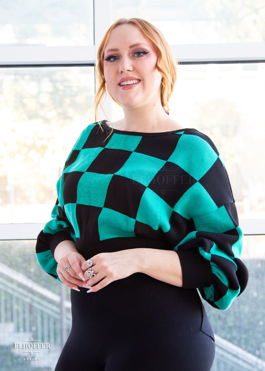 Kelsey, a fair skinned S model with strawberry blonde hair, is smiling while wearing the XL/2XL sample of a cropped oversized sweater with a black and green chessboard pattern and long billowing sleeves.  She would normally wear the XS/S.  She paired the sweater with black leggings.