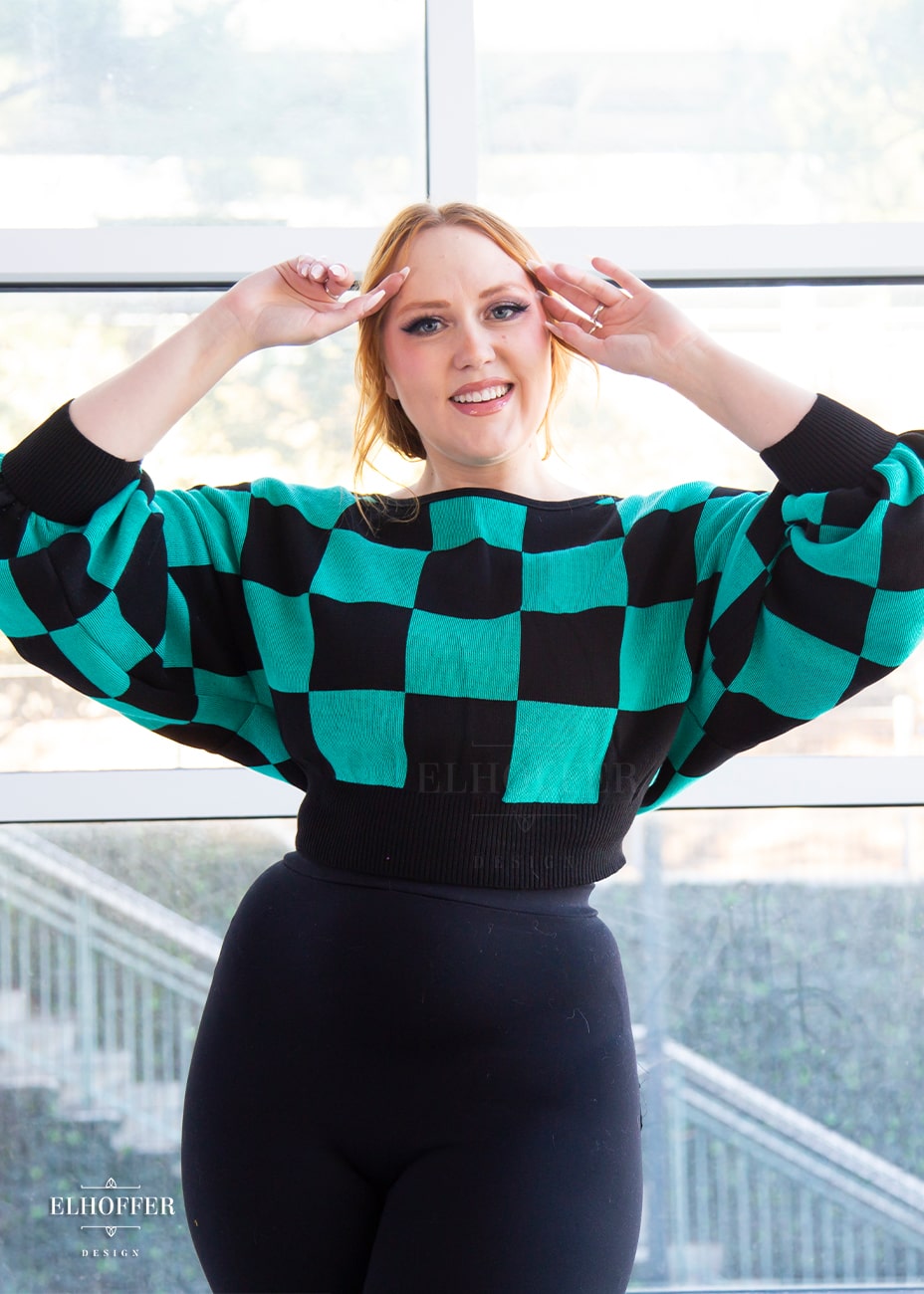 Kelsey, a fair skinned S model with strawberry blonde hair, is smiling while wearing the XL/2XL sample of a cropped oversized sweater with a black and green chessboard pattern and long billowing sleeves.  She would normally wear the XS/S.  She paired the sweater with black leggings.