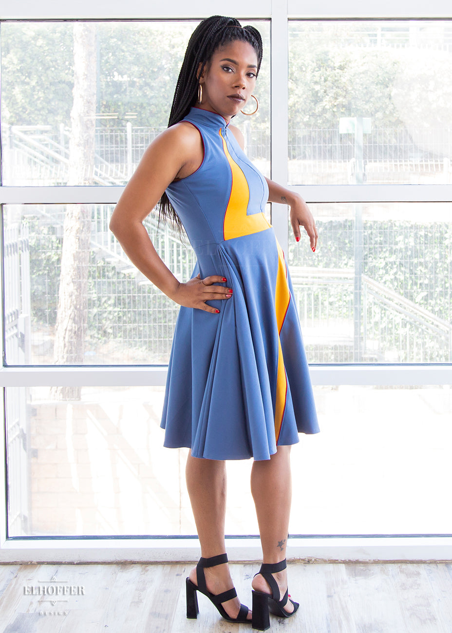 Krystina, a size small medium dark skinned model with long braids, shows of the side of the high-necked sleeveless blue dress. The dress is piped in crimson and has a yellow lightening bolt emblazoned over the front of it. There is a zip down the middle down to the waist.