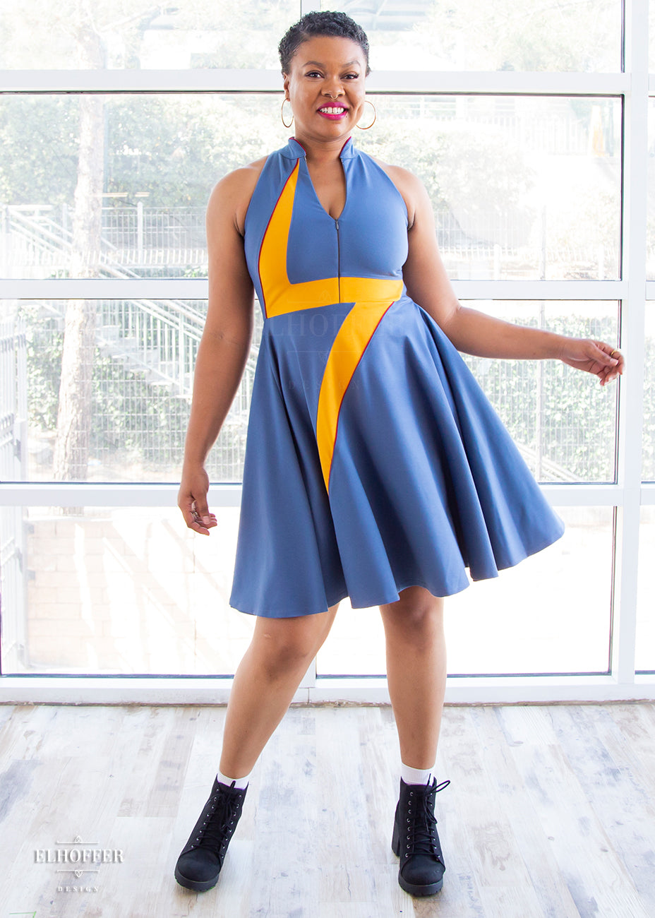 Desiree, a size large medium dark skinned model with very short dark hair, wears a high-necked sleeveless blue dress. The dress is piped in crimson and has a yellow lightening bolt emblazoned over the front of it. There is a zip down the middle down to the waist that she has unzipped to show a bit of décolletage.