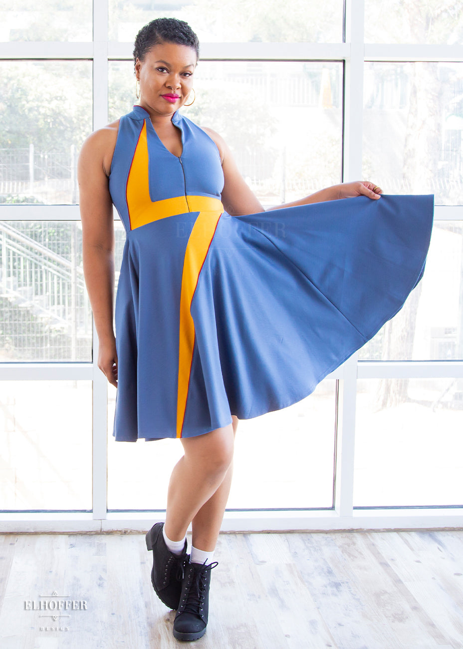 Desiree, a size large medium dark skinned model with very short dark hair, holds out the full skirt of a high-necked sleeveless blue dress. The dress is piped in crimson and has a yellow lightening bolt emblazoned over the front of it. There is a zip down the middle down to the waist that she has unzipped to show a bit of décolletage.