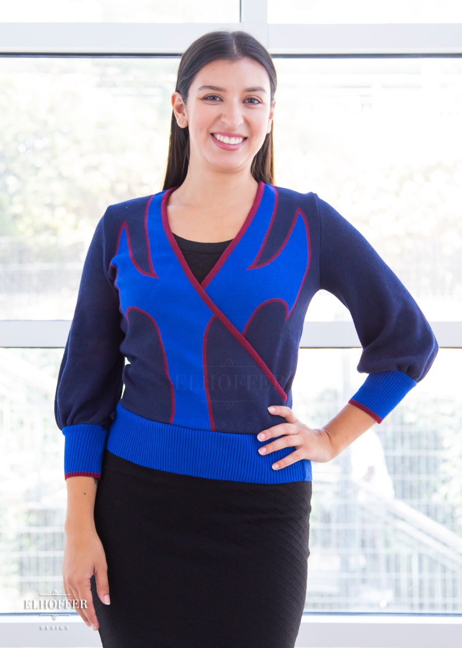 Alexandra, an olive skinned size small model with long brown hair, is wearing a crossover knit crop top. The base is a dark blue, details are bright blue with a red outline and form almost a t shape in the middle of the crop. The cuffs and bottom are the same bright blue of the detail and the crossover is piped in the same red.