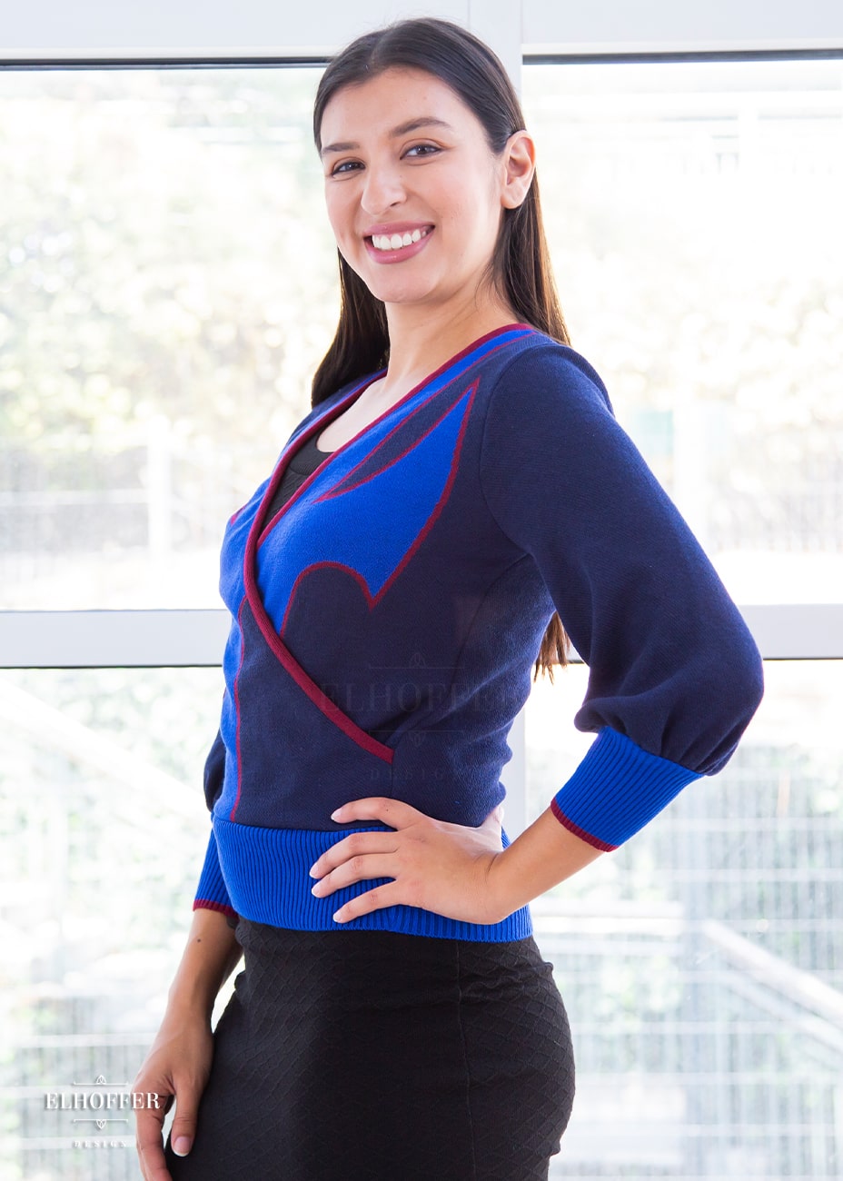 Alexandra, an olive skinned size small model with long brown hair, is wearing a crossover knit crop top. The base is a dark blue, details are bright blue with a red outline and form almost a t shape in the middle of the crop. The cuffs and bottom are the same bright blue of the detail and the crossover is piped in the same red.