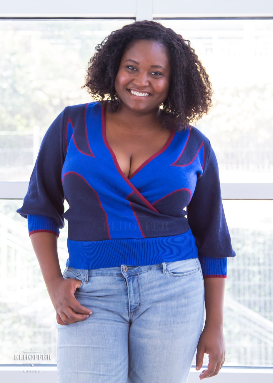 Maydelle, a medium dark skinned size XL model with shoulder length curly brown hair, is wearing a crossover knit crop top. The base is a dark blue, details are bright blue with a red outline and form almost a t shape in the middle of the crop. The cuffs and bottom are the same bright blue of the detail and the crossover is piped in the same red.