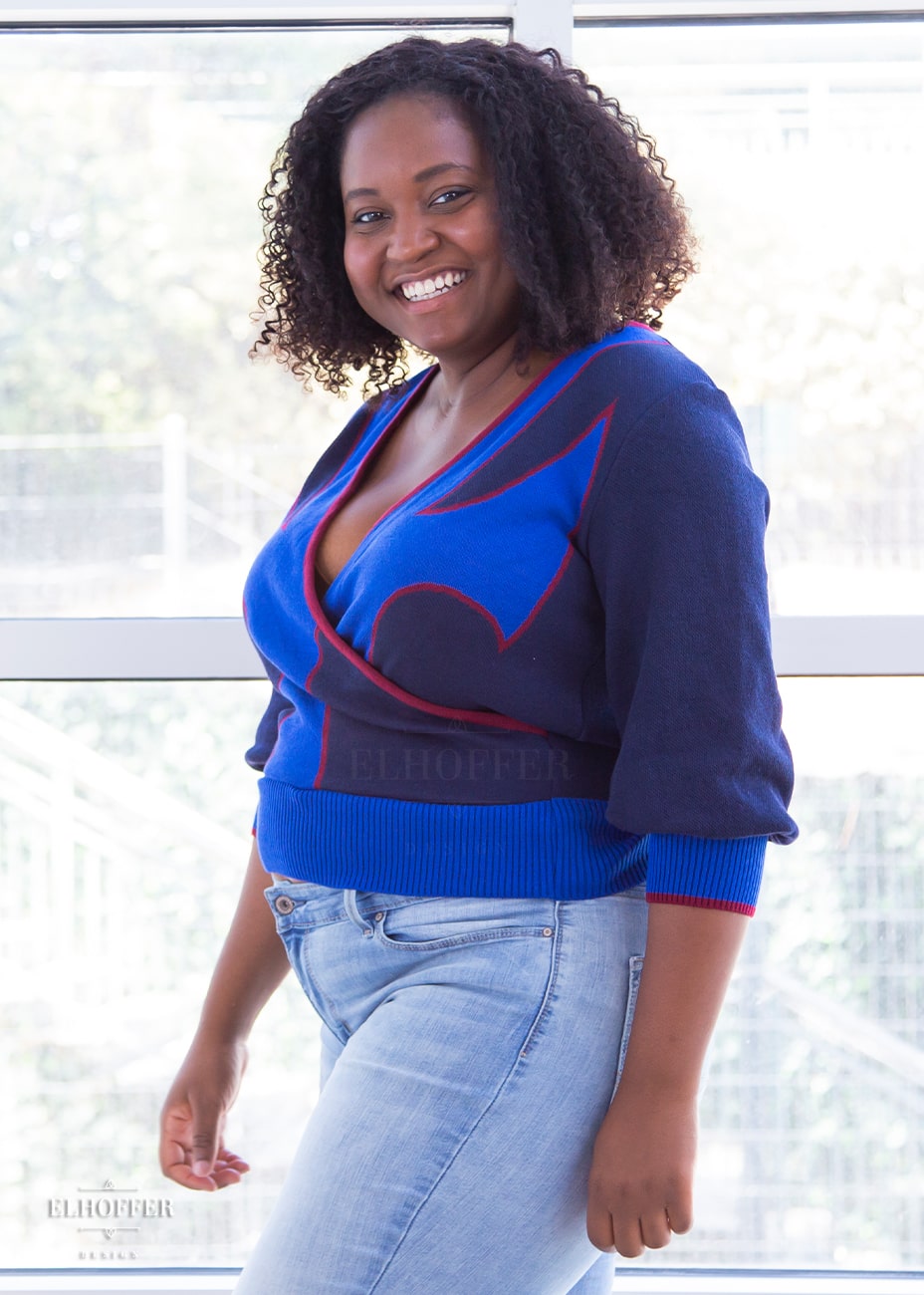 Maydelle, a medium dark skinned size XL model with shoulder length curly brown hair, is wearing a crossover knit crop top. The base is a dark blue, details are bright blue with a red outline and form almost a t shape in the middle of the crop. The cuffs and bottom are the same bright blue of the detail and the crossover is piped in the same red.
