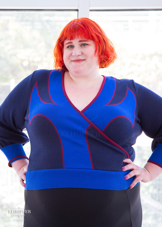 Logan, a fair skinned size 3XL model with bright orange hair and bangs, is wearing a crossover knit crop top. The base is a dark blue, details are bright blue with a red outline and form almost a t shape in the middle of the crop. The cuffs and bottom are the same bright blue of the detail and the crossover is piped in the same red.